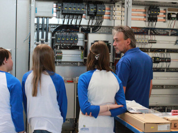 Ein Mitarbeiter zeigt drei Mädels die Komponenten eines Schaltschrankes