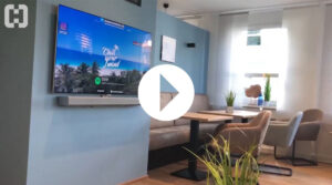 Einblick in die Business Lounge: EIn TV an der Wand mit Tischen und Stühlen - Link führt zum YouTube-Video