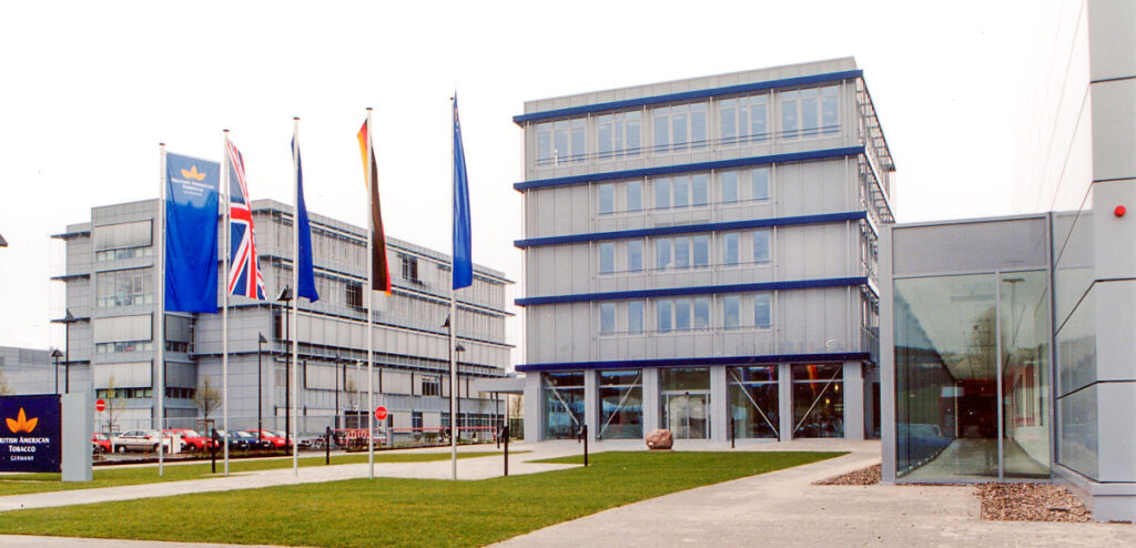 Außenansicht des Firmengebäudes „British American Tobacco Bayreuth“ mit zwei Gebäudekomplexen und verschiedenen Länderflaggen davor
