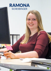 Blonde Frau mit weinrotem Shirt sitzt an einem PC und lacht in die Kamera