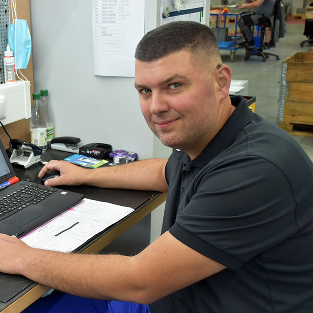 A man with black shirt and shaved hair sits at a workbench with a laptop in front of him