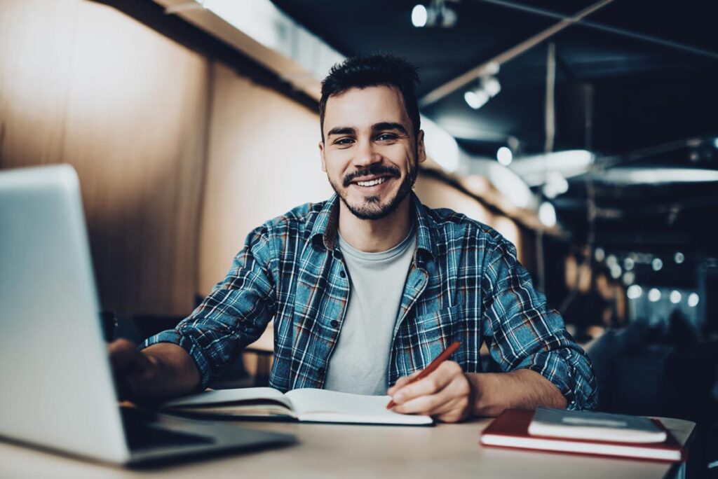 Junge sitzt in Cafe vor Laptop und macht sich Notizen