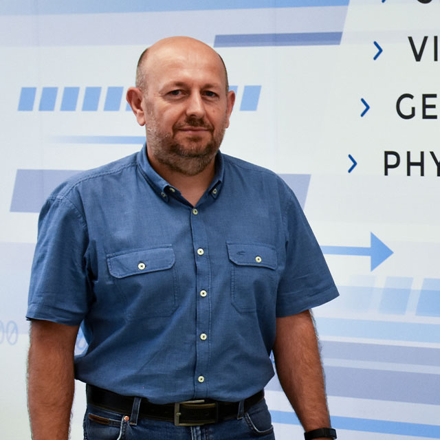 A man with half bald head, blue shirt and jeans stands in front of a white wall with blue arrows