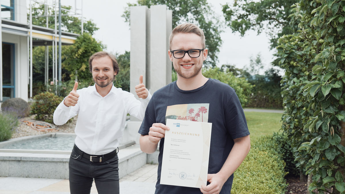 Ein junger Mann mit kurzen blonden Haaren und Brille steht mit einer Urkunde vor einem Brunnen. Im Hintergrund zeigt ein zweiter Mann mit braunen Haaren beide Daumen nach oben