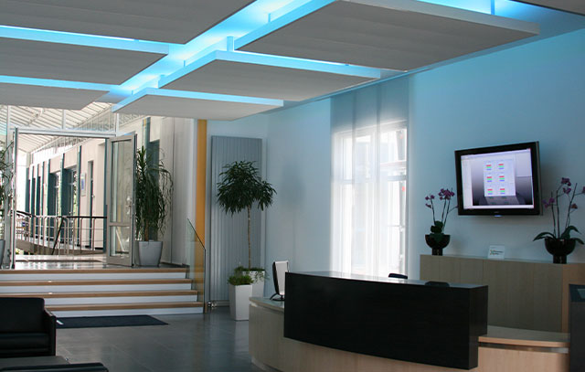 Entrance hall with reception desk and TV on the wall - on the ceiling hang 6 white squares that shine light blue on the edge