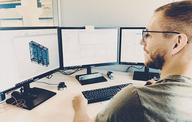 Ein Mann mit Brille und grünem Shirt sitzt am Schreibtisch mit drei Bildschirmen – auf einem Bildschirm ist ein 3D-Modell eines Schaltschrankes zu sehen