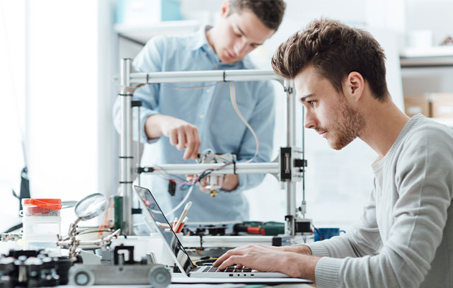 A man sits at a laptop, in the background another man stands at a 3D printer