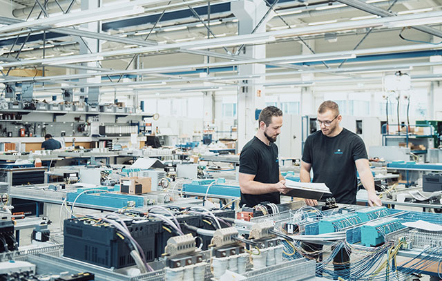 Two men stand with a folder in their hands in the production hall in front of many assembly plates laid on tables