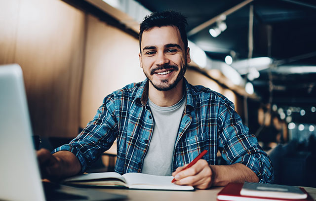 A young man sits in front of a laptop with a book