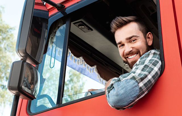 A man with brown hair and check shirt sits in a red truck and smiles out the open window