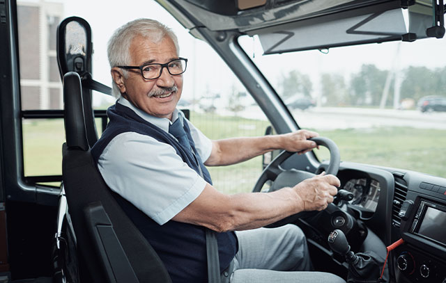 Ein Mann Sitzt an einem Lenkrad in einem kleinen Bus und lächelt in die Kamera