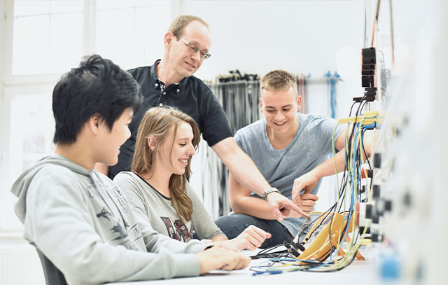 3 young trainees sit at a workbench and let their instructor explain something to them