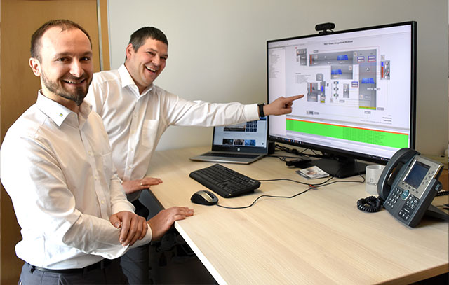 Zwei HERMOS-Mitarbeiter stehen an einem höhenverstellbarem Schreibtisch und zeigen auf einem großen Monitor mit einer FIS Applikation