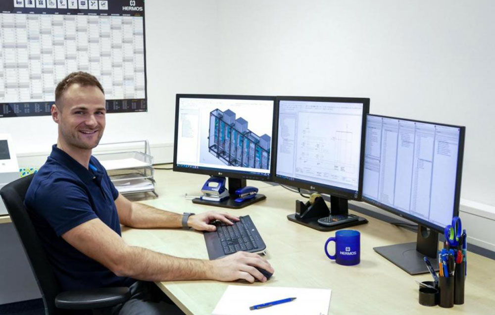 A man sits in front of 3 screens at a desk